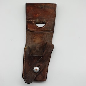 May include: A brown leather holster with a loop at the top and a flap secured by a silver snap. The holster shows visible stitching and a worn appearance, suggesting age and use. The leather is a rich, dark brown colour.