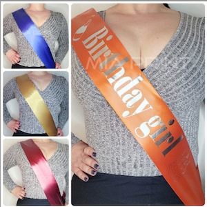 Könnte beinhalten: Orangener Satin-Schärpe mit silbernem Text "Birthday Girl" und einer Champagnerflaschen-Grafik.