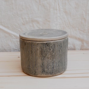 May include: A cylindrical ceramic container with a lid. The container is light beige with vertical black lines and a circular pattern on the lid. The container is sitting on a light wood surface.