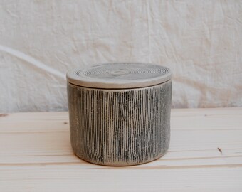 May include: A cylindrical ceramic container with a lid. The container is light beige with vertical black lines and a circular pattern on the lid. The container is sitting on a light wood surface.