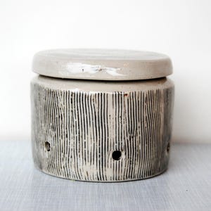 Può includere: Vaso in ceramica grigio chiaro con coperchio, superficie strutturata e linee nere verticali. Il vaso cilindrico ha tre piccoli fori. Ideale per la conservazione in cucina, alto circa 15 cm.