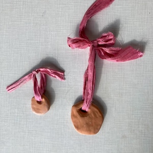 May include: Two terracotta clay pendants with pink fabric loops. The larger pendant is round and the smaller pendant is oval.