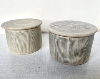 May include: Two cylindrical ceramic containers with matching lids. Each piece features a light beige base colour with dark grey vertical etched lines on the sides and a circular, concentric ring pattern engraved on the top of the flat lids.