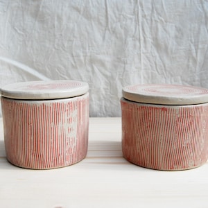 May include: Two ceramic jars with lids. The jars are cylindrical with a cream-colored base and vertical red stripes. The lids are cream-colored with a spiral design. The jars are on a light wooden surface.
