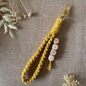 Puede incluir: Un llavero de macramé amarillo mostaza con un cierre de metal dorado y cuatro cuentas de madera que deletrean el nombre "KIKI".