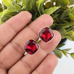 Puede incluir: Un par de gemelos de plata con piedras rojas facetadas de corte cuadrado. Las piedras tienen una superficie pulida y reflectante, engastadas en un marco de metal plateado sencillo con un diseño de bordes suaves y redondeados.