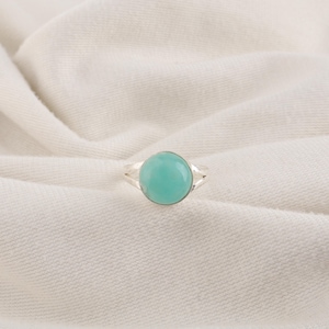 Peut inclure: Une bague en argent ornée d'une pierre cabochon ronde et lisse de couleur turquoise clair, sertie dans une monture métallique simple. L'anneau de la bague est divisé en deux fins fils d'argent polis qui se rejoignent à la base du sertissage.