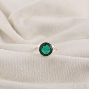 May include: A silver ring featuring a round, faceted, vibrant green gemstone set in a simple metal band. The stone displays a clear, brilliant cut with multiple facets that reflect light, creating a bright and saturated emerald-like appearance.