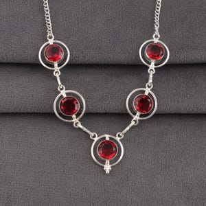 May include: A silver necklace featuring five circular links, each containing a round, faceted deep red gemstone. The links are connected by small silver rings and a delicate silver chain, creating a symmetrical, open-work design.