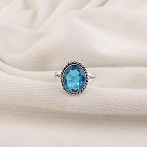 Puede incluir: Un anillo de plata con una gran piedra preciosa de color azul brillante, de corte ovalado, engastada en un borde de metal con diseño de cuerda trenzada. El aro del anillo se divide en dos segmentos finos de plata pulida que se unen.