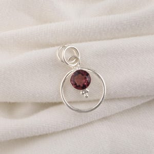 May include: A silver pendant featuring a circular frame with a round, deep red faceted gemstone suspended in the center. The gemstone is accented by three small silver beads at its base, and the pendant includes a double silver jump ring bail.
