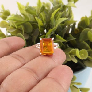 May include: A silver ring featuring a large, rectangular, faceted orange gemstone set in a four-prong mounting. The band is split into two thin silver segments that meet at the base of the stone setting, creating a clean and polished appearance.