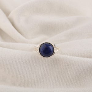 以下が含まれることがあります： 中央に滑らかで丸い深い青色のラピスラズリの宝石をあしらったシルバーリング。リングのバンドは磨かれたシルバーで作られており、2本の細く繊細なアームに分かれて、円形の石のセッティングの基部で合流しています。