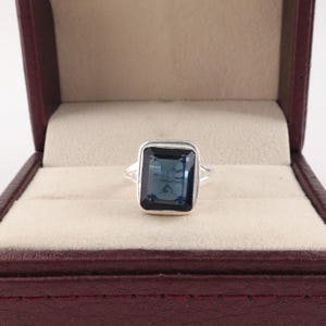 May include: A silver ring featuring a large, rectangular, faceted dark blue gemstone set in a smooth metal bezel. The band of the ring is split into two thin, polished silver segments that meet at the base of the central stone setting.