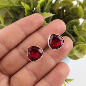 Puede incluir: Un par de pendientes de botón con gemas facetadas de color rojo intenso y forma triangular, engastadas en metal pulido de tono plateado. Las piedras tienen un corte brillante que refleja la luz, creando un aspecto vibrante.