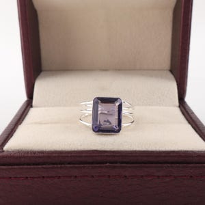 May include: A silver ring featuring a large, rectangular, faceted purple gemstone set in a prong setting. The band consists of three thin, parallel silver wires that converge at the base of the stone, creating a layered, open-work design.
