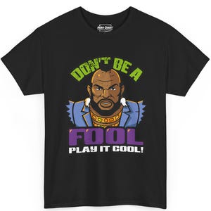 May include: Black t-shirt featuring a cartoon illustration of a stern-faced man with a gold chain and the text "DON'T BE A FOOL PLAY IT COOL!" in green and purple. The shirt has a crew neck and short sleeves.