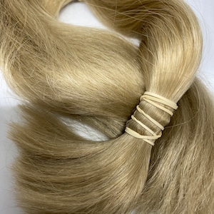 May include: Two bundles of light blonde hair extensions, secured with beige rubber bands. The hair is a natural, wavy texture, and the color is a warm, neutral blonde. The bundles are arranged in a slightly overlapping pattern.