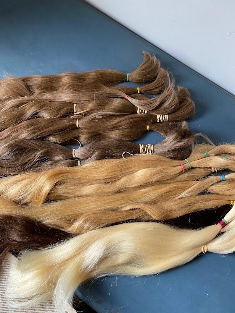 May include: Several bundles of hair extensions in various shades of brown and blonde are arranged on a blue surface. Each bundle is secured with a small rubber band. The hair colors range from dark brown to light blonde.