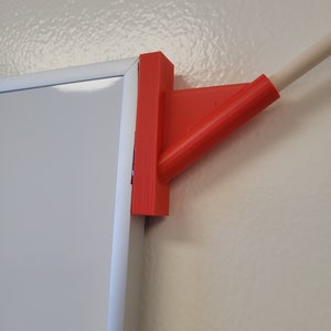 May include: A red plastic corner bracket for mounting a whiteboard to a wall. The bracket is attached to the whiteboard frame and has a hole for a screw.