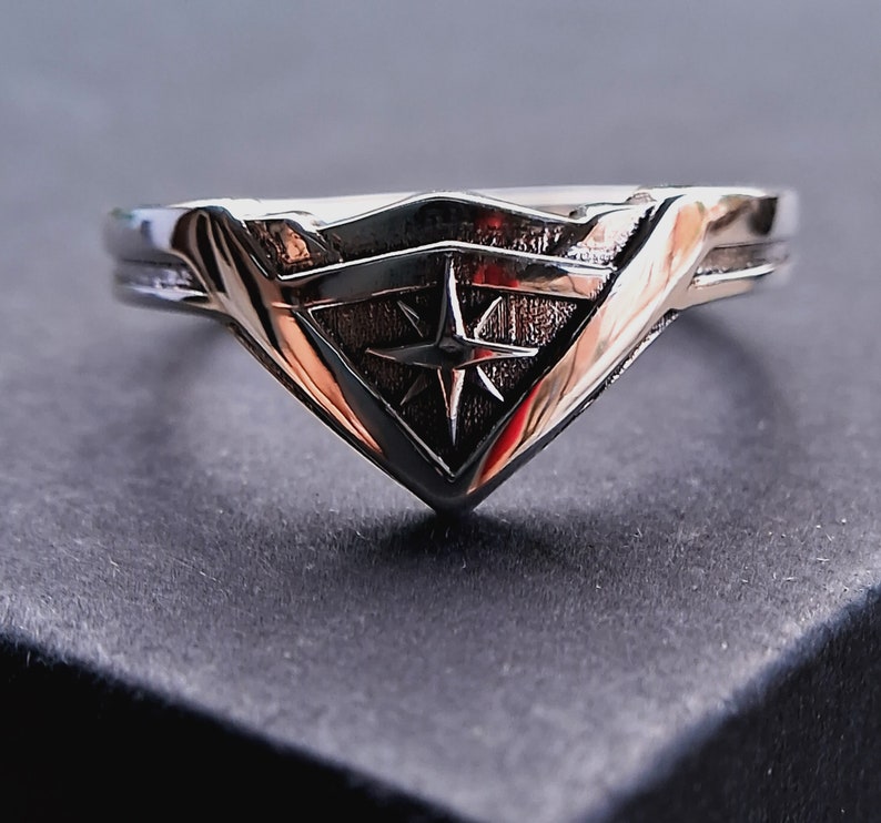 Pu&ograve; includere: Un anello d'argento con un design a stella stilizzato all'interno di una forma a scudo.