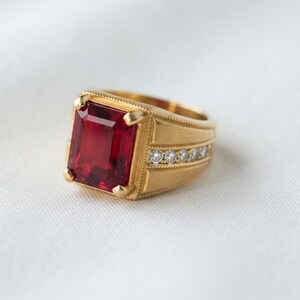 May include: A gold ring featuring a large, rectangular, emerald-cut red gemstone set in the center. The band has a brushed matte finish and is accented with a row of small, round, clear white stones embedded along the side of the shank.