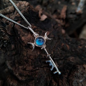 May include: A silver pendant necklace with a key design. The key features a central blue gemstone, framed by crescent moons. The key's handle and teeth are also visible. The necklace chain is a delicate, linked design.