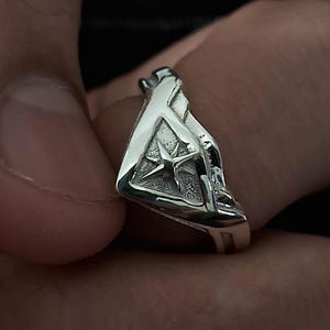Pu&ograve; includere: Un anello d'argento con un design a forma di stella al centro. L'anello ha la forma di un triangolo con una superficie testurizzata.
