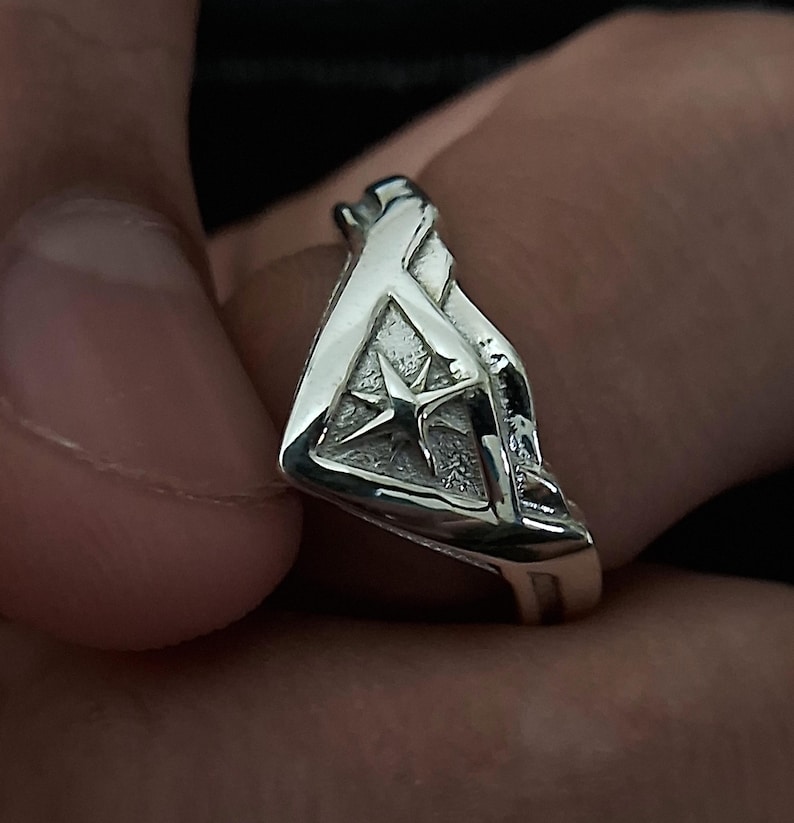 Pu&ograve; includere: Un anello d'argento con un design a forma di stella al centro. L'anello ha la forma di un triangolo con una superficie testurizzata.