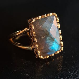 May include: A gold ring featuring a large, rectangular, iridescent labradorite gemstone set in a gold bezel. The ring has a simple, open band.