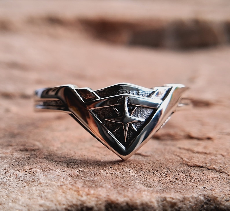Pu&ograve; includere: Un anello d'argento con un design a stella stilizzato al centro. L'anello ha la forma di uno scudo con la parte inferiore a punta.