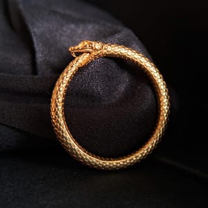 May include: A gold snake ring with a detailed snake head design. The ring is on a black velvet background.