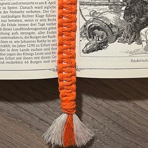 Op de afbeelding: Een close-up van een oranje en witte gehaakte bladwijzer met een kwast. De bladwijzer ligt op een open boek met tekst over een historische gebeurtenis.