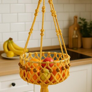 May include: A hanging macrame fruit basket in a mustard yellow color, filled with oranges, apples, and a green apple. The basket is adorned with wooden beads and a tassel. Bananas are visible in the background.