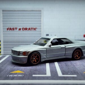 May include: A silver toy car model of a Mercedes-Benz 560 SEC AMG with a black interior and gold wheels. The car is parked in a garage setting with a sign that reads "FAST & DRATIC".