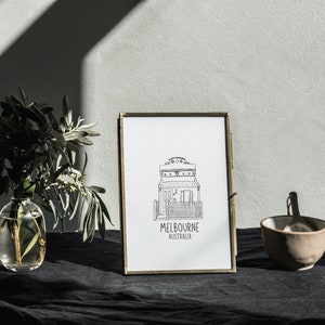 May include: A black and white line drawing of a house with the text "MELBOURNE AUSTRALIA" underneath, framed in a gold frame. The frame is sitting on a black surface with a vase of greenery and a small bowl to the right.