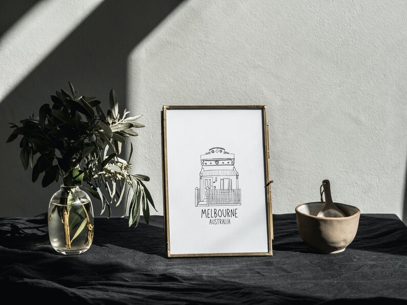 May include: A black and white line drawing of a house with the text "MELBOURNE AUSTRALIA" underneath, framed in a gold frame. The frame is sitting on a black surface with a vase of greenery and a small bowl to the right.