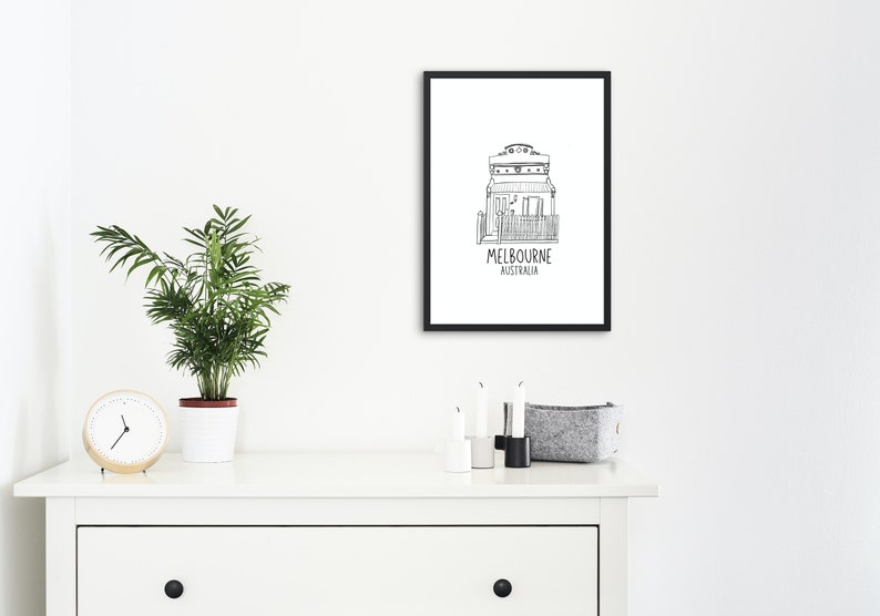 May include: Black and white line drawing of a house with the text "MELBOURNE AUSTRALIA" underneath. The drawing is framed in a black frame and hanging on a white wall.