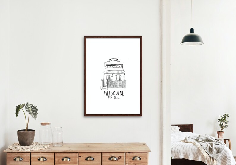May include: Black and white line drawing of a house with the text "MELBOURNE AUSTRALIA" underneath. The drawing is framed in a brown frame and is hanging on a white wall.