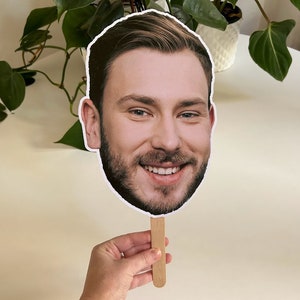 May include: A cardboard cutout of a man's face with a beard and a smile, attached to a wooden stick. The cutout is held in a hand.