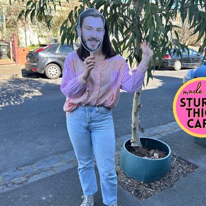 May include: A person holds a cardboard cutout of a man's face in front of their face. The cutout is on a stick. The person is wearing a pink and white striped shirt and blue jeans. The person is standing in front of a tree and a planter. The text "made from STURDY THICK CARD" is on a pink circle.