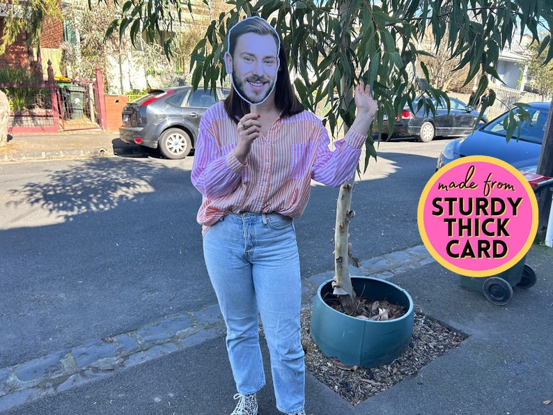May include: A person holds a cardboard cutout of a man's face in front of their face. The cutout is on a stick. The person is wearing a pink and white striped shirt and blue jeans. The person is standing in front of a tree and a planter. The text "made from STURDY THICK CARD" is on a pink circle.