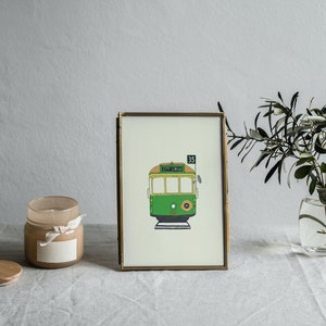 May include: A gold framed print of a green and yellow tram with the number 35 and the text "CITY CIRCLE" on the front. The print is on a white background and is sitting on a white table with a candle and a vase of greenery.