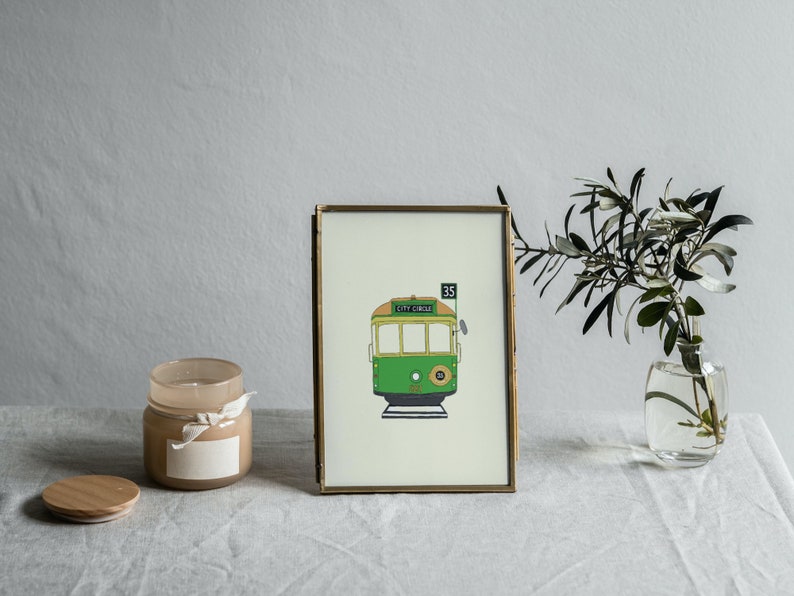 May include: A gold framed print of a green and yellow tram with the number 35 and the text "CITY CIRCLE" on the front. The print is on a white background and is sitting on a white table with a candle and a vase of greenery.