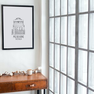 May include: Black and white line drawing of a house with the text "MELBOURNE AUSTRALIA" underneath. The drawing is framed and hanging on a white wall. A wooden table with two drawers is in the foreground.