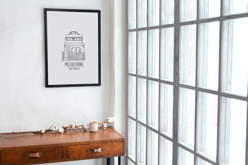 May include: Black and white line drawing of a house with the text "MELBOURNE AUSTRALIA" underneath. The drawing is framed and hanging on a white wall. A wooden table with two drawers is in the foreground.