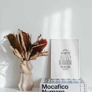 May include: A white vase with a wavy design holds a bouquet of dried brown and red flowers. A white framed print with a black and white illustration of a house and the text "Melbourne Australia" sits on a white surface next to a stack of books with the text "Mocacifico Numero" in black.