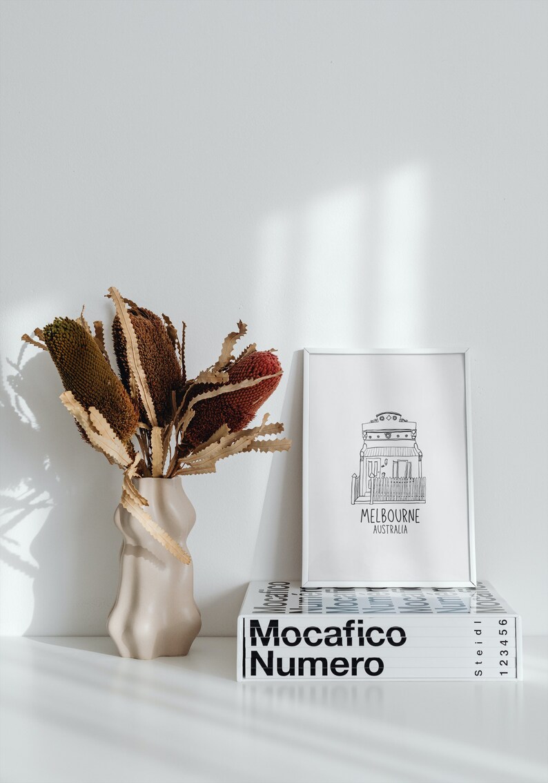May include: A white vase with a wavy design holds a bouquet of dried brown and red flowers. A white framed print with a black and white illustration of a house and the text "Melbourne Australia" sits on a white surface next to a stack of books with the text "Mocacifico Numero" in black.