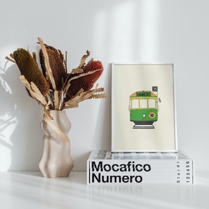 May include: A framed illustration of a green and yellow tram with the number 35 and the words "City Circle" on the front. The frame is white and is sitting on top of a stack of books with the word "Mocacifico Numero" printed on the front.