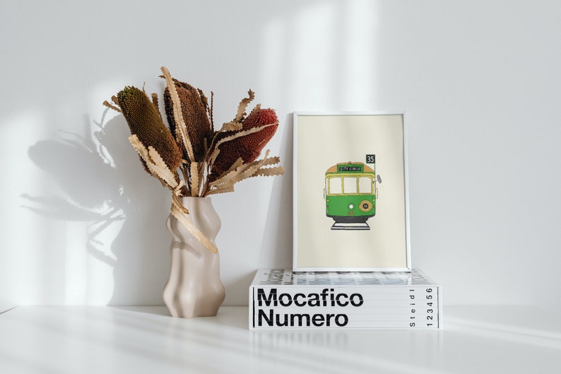 May include: A framed illustration of a green and yellow tram with the number 35 and the words "City Circle" on the front. The frame is white and is sitting on top of a stack of books with the word "Mocacifico Numero" printed on the front.